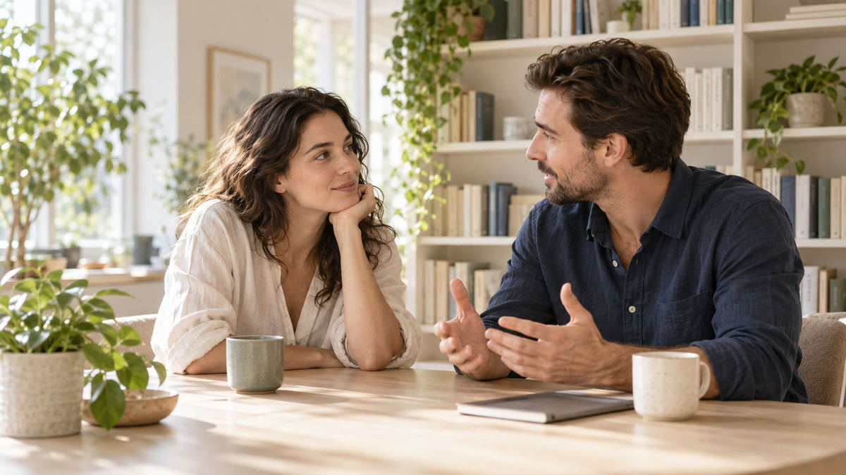 Two adults talking at a pale wooden table in a sunlit library